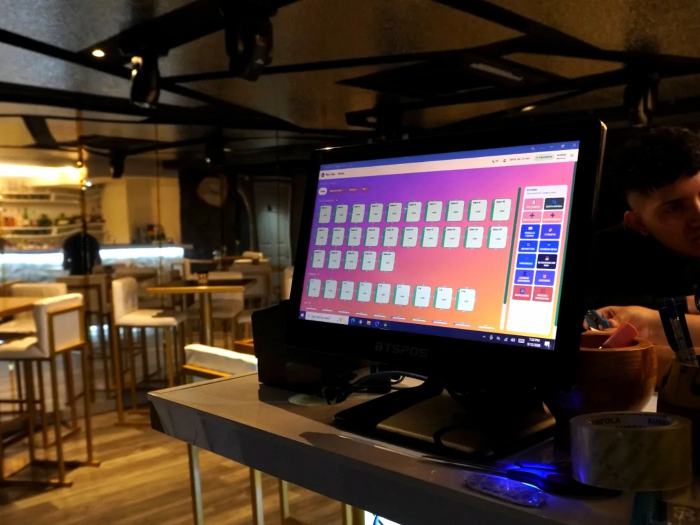Monitor táctil sobre la barra de un restaurante mostrando el mapa de mesas de SuiteHub, con el salón de mesas altas al fondo.