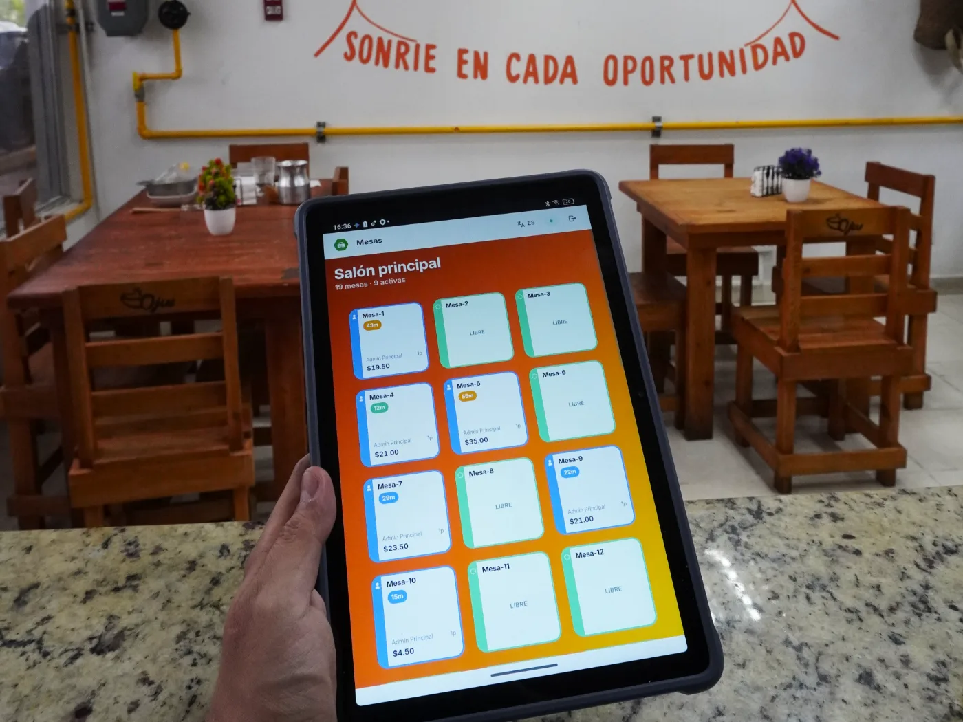 Tablet con HubPOS mostrando las mesas activas del salón de Ojueee, con el comedor de fondo