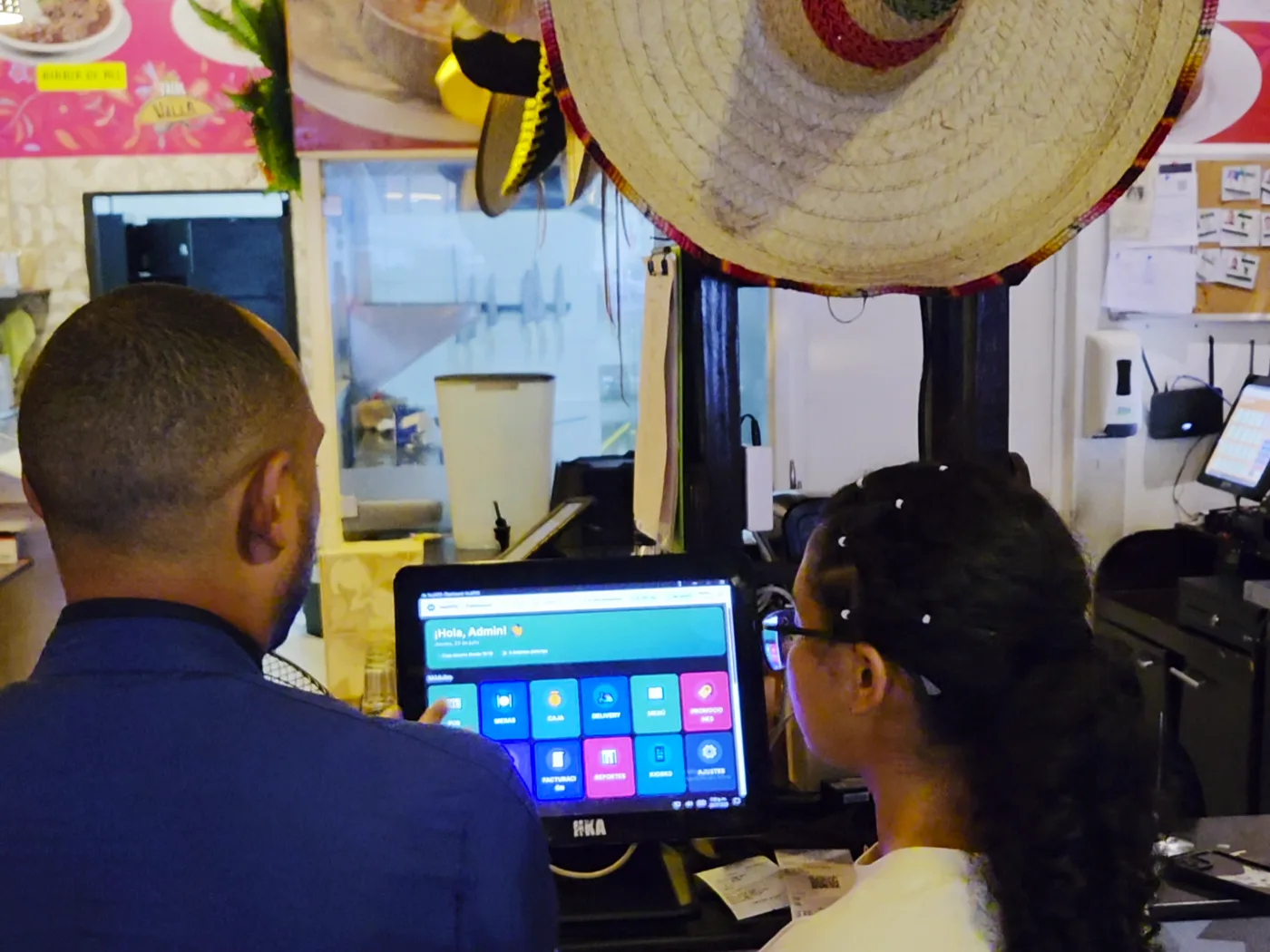 Dos personas del equipo de Los Tacos de Villa frente al tablero de inicio de HubPOS en el mostrador