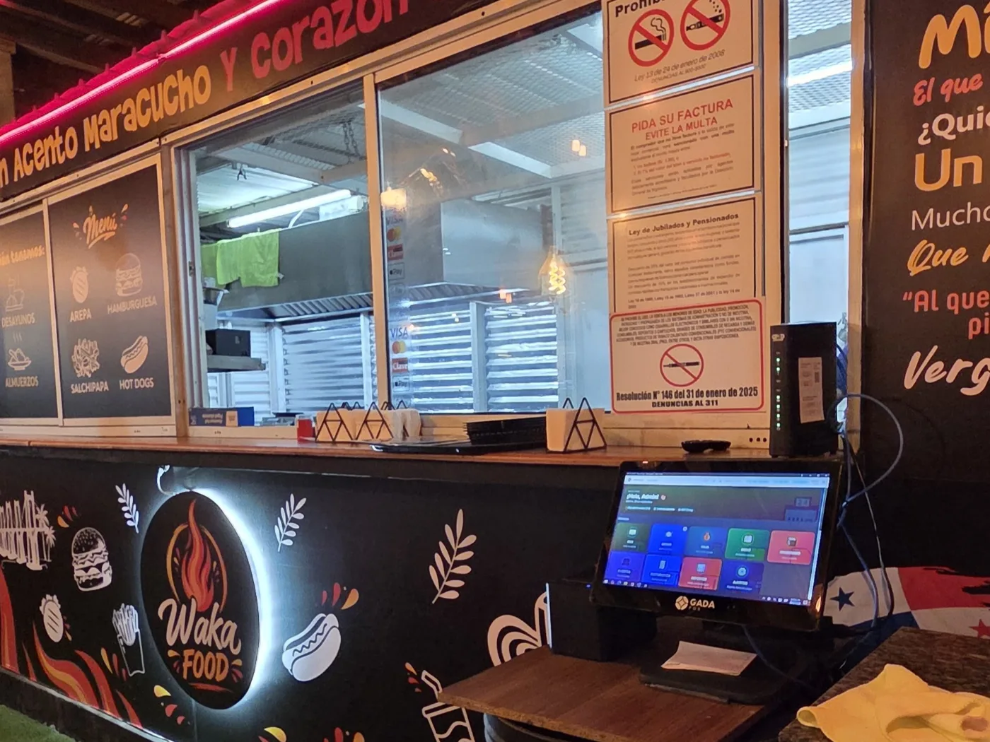 Barra de un local de comida rápida con menú iluminado y una pantalla táctil de punto de venta sobre el mostrador.